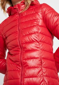 Chaqueta de plumas roja con capucha, cierre de cremallera, diseño acolchado y acabado brillante. Acabada con un sutil logo de la marca en la manga.