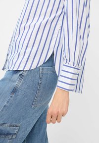 Blauw-wit gestreepte blouse met lange mouwen en een gebogen zoom, voorzien van knoopsluitingen bij de mouwen en gemaakt van een zachte, lichte stof. Gepaard met denim.