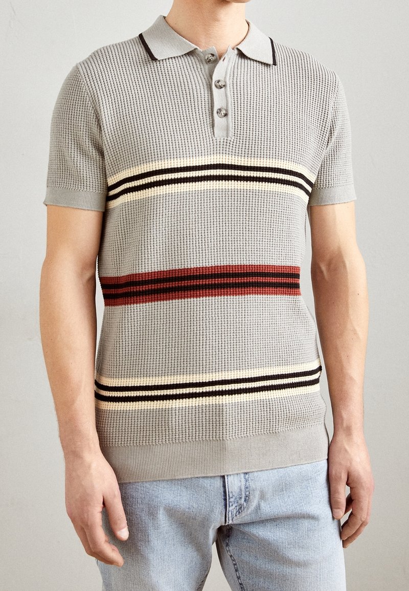 Polo de color gris con textura que presenta tres franjas horizontales en rojo, crema y negro. Mangas cortas y un cuello con tres botones.