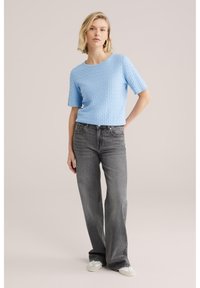 Vaaleansininen teksturoitu lyhyt neule, jossa on lyhyet hihat, yhdistetty korkea vyötäröisiin harmaisiin levenevillä lahkeilla varustettuihin farkkuihin ja vaaleisiin kenkiin.