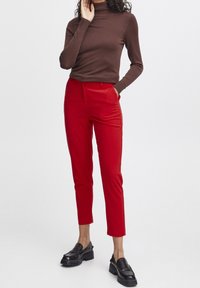 Rode op maat gemaakte broek met een gladde textuur, voorzien van een platte voorkant en zijzakken. Draagt bij een aangesloten bruine coltrui en zwarte schoenen.
