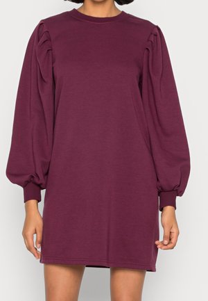 Femme portant une robe prune à manches longues avec des épaules bouffantes et des poignets, debout devant un fond clair uni.