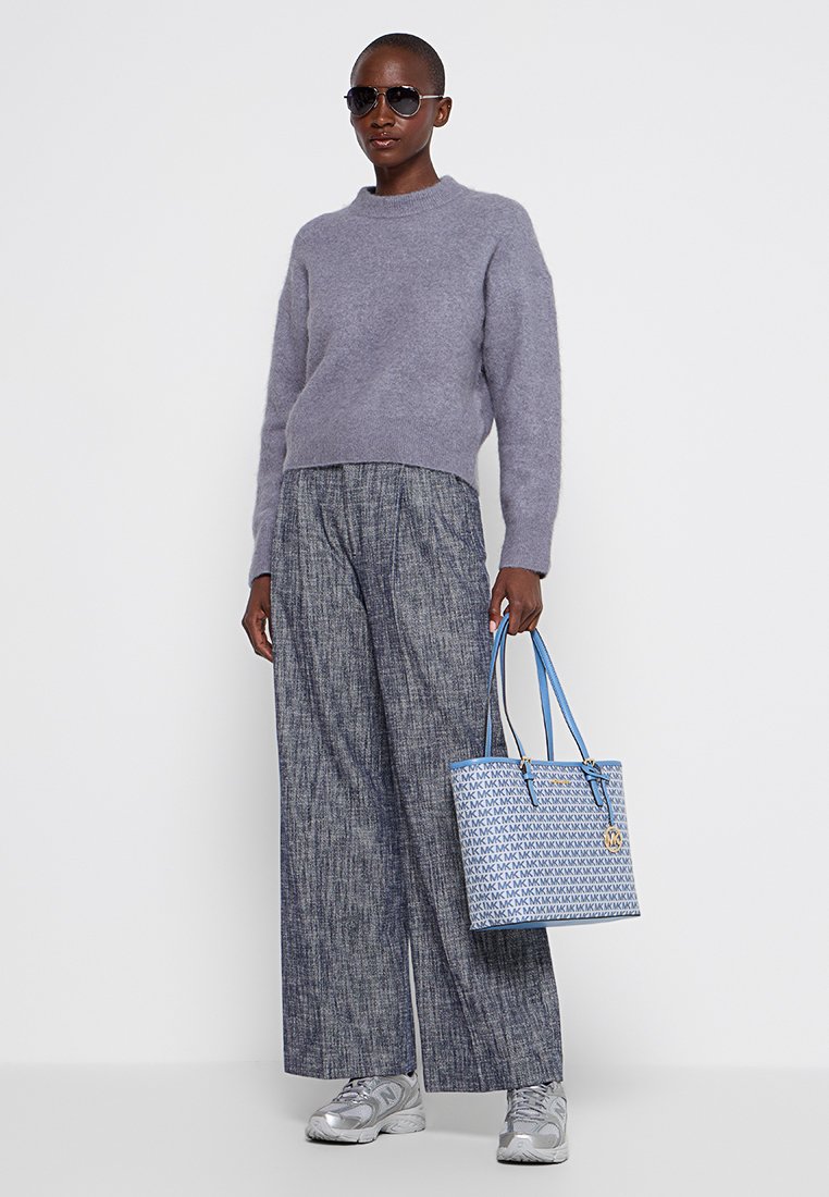 Maglione grigio chiaro, pantaloni a quadri larghi e una borsa blu con motivo. La modella indossa occhiali da sole e sneaker argentate.