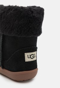 Bottine en suede noir avec un bord en laine de mouton noir et moelleux, semelle en caoutchouc et étiquette UGG cousue sur le côté. Présente une finition texturée.