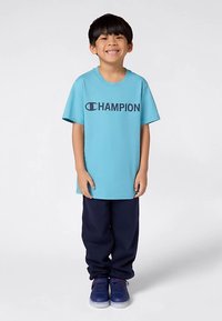 Χαμογελαστό παιδί που στέκεται, φορώντας ελαφρύ μπλε T-shirt Champion, σκούρα μπλε παντελόνια και μπλε αθλητικά παπούτσια σε απλό ανοιχτό φόντο.