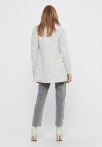 Cappotto lungo bianco maculato con un design strutturato, abbinato a pantaloni dritti grigi sbiaditi e stivaletti crema alla caviglia. Look semplice e moderno.