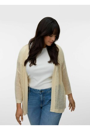 Žena s dlhými tmavými vlasmi, ktorá má na sebe krémový pletený sveter, bielu blúzku a modré džínsy, pozerá sa dole s uvoľnenými rukami.