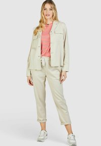 Lichtbeige linnen jasje met twee borstzakken over een rood-wit gestreept T-shirt. Bijpassende beige broek met trekkoord en grijze sneakers.