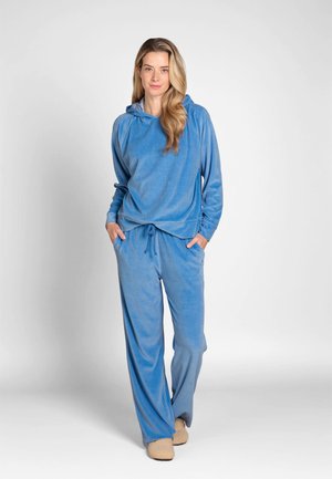 Woman with long blonde hair wearing a blue hooded sweatshirt and matching loose pants, standing with hands in pockets on a plain background.