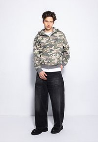 Bluza z kapturem w kamuflażu w odcieniach zieleni i beżu, z szarym wykończeniem i logo. Połączona z luźnymi, czarnymi dżinsami i czarnymi butami.