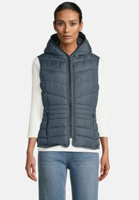 Vrouw in een mouwloze marineblauwe gewatteerde vest met capuchon over een witte langemouwige geribbelde shirt en blauwe jeans, die recht vooruit kijkt met ontspannen armen.