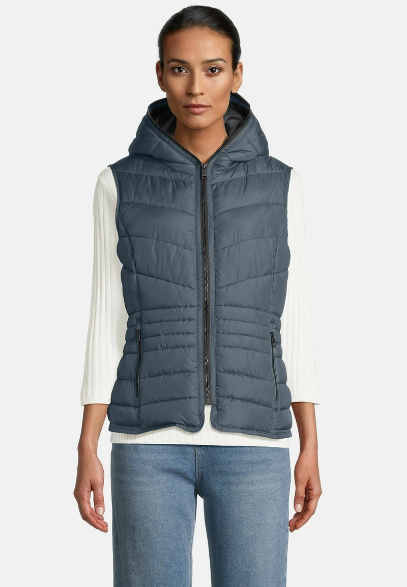 Vrouw in een mouwloze marineblauwe gewatteerde vest met capuchon over een witte langemouwige geribbelde shirt en blauwe jeans, die recht vooruit kijkt met ontspannen armen.