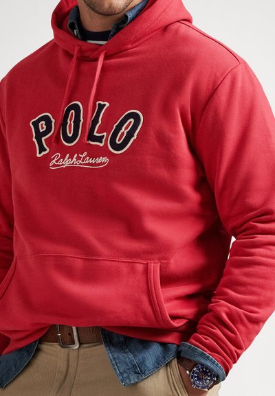 Άντρας που φοράει κόκκινο φούτερ Polo Ralph Lauren, μπλε πουκάμισο από τζιν από κάτω, μπεζ παντελόνι, καφέ ζώνη και μπλε ρολόι χειρός.