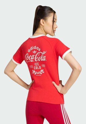 Kvinde med mørkt hår i hestehale iført en rød Adidas x Coca-Cola T-shirt og røde Adidas bukser med hvide striber, hænderne på hofterne, set fra siden.