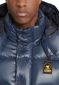 Giacca imbottita navy in nylon lucido, collo alto, bottoni a pressione, con una toppa logo nera e gialla sul davanti.