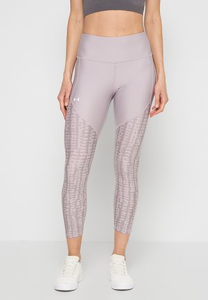 Femme portant des leggings violet clair à motifs et des baskets blanches, debout devant un fond clair uni.