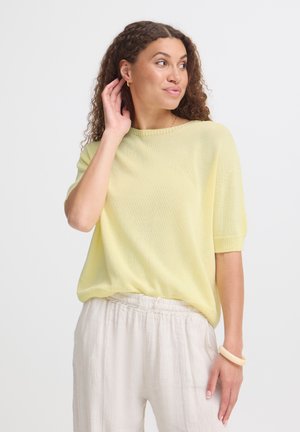 Kvinde med krøllet hår iført lysegul kortærmet sweater, hvide løse bukser, gyldne creoløreringe og beige armbånd, kigger til siden.
