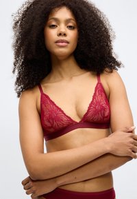 Crvena čipkasta bralette s dizajnom u obliku V, mrežastim detaljima i mekanim potporom ispod. Ima podesive naramenice i cvjetni uzorak.