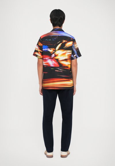 Camicia a maniche corte con un vivace motivo astratto nei toni del rosso, blu e giallo su sfondo nero. Abbinata a pantaloni blu navy.