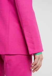Bras droit et main d'une personne portant une veste de costume rose vif et un pantalon assorti, avec un poignet de chemise vert visible.