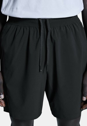 Sorte elastiske shorts med snøre i taljen, båret af en person med mørk hud, kombineret med en hvid indstoppet skjorte.