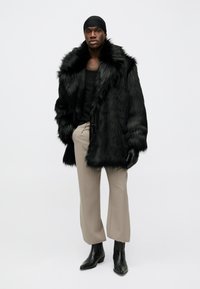Black faux fur coat with a large collar, paired with beige trousers and black ankle boots. Accessories include a black head wrap and gloves.