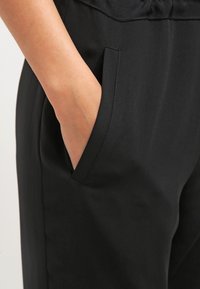 Svarta byxor med en sidoficka, tillverkade av ett slätt tyg med en lätt glans. Midjebandet är elastiskt för extra komfort.