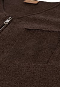 Rudas vilnonis kardiganas su užtrauktuku, krūtinės kišene ir švelnia, tekstūrine medžiaga. Dizainas minimalistinis, su švariomis linijomis.