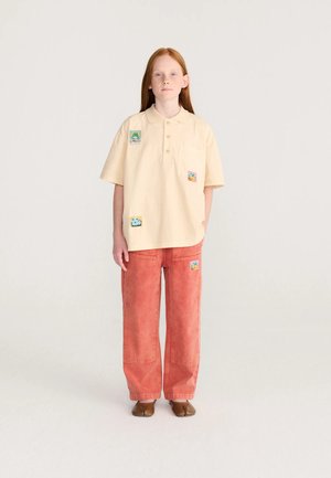Barn med langt rødt hår står iført en beige polotrøje med farverige lapper og løse røde bukser med en lap på låret, brune sko.