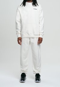Felpa bianca in cotone con tasca frontale e logo, abbinata a pantaloni della tuta bianchi coordinati e scarpe da ginnastica nere. Texture liscia, vestibilità comoda.