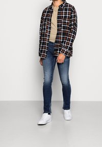 Ung person klädd i en svart och orange rutad skjorta över en beige t-shirt, blå tajta jeans och vita sneakers, står mot en enfärgad bakgrund.