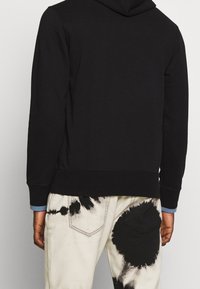Felpa nera con una texture liscia, dotata di tasca a canguro e cappuccio regolabile con coulisse. Abbinata a pantaloni chiari con motivo tie-dye.