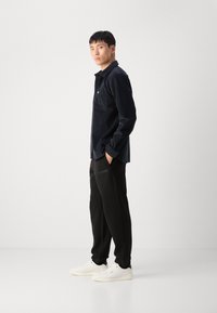 Chemise en velours côtelé bleu marine à boutons avec une poche sur la poitrine, associée à un pantalon de survêtement noir avec une bande texturée et des baskets blanches.