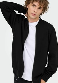 Sort zip-up sweatshirt med ribbet tekstur, rund halsudskæring og lange ærmer, båret over en hvid T-shirt. Sølv lynlåsdetalje.