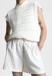 Top in cotone all'uncinetto bianco con collo rotondo e maniche corte, sovrapposto a pantaloni corti bianchi con cordoncino che presentano un motivo sottile.