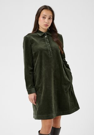Femme portant une robe en velours côtelé vert foncé à manches longues avec des boutons, debout avec une main dans la poche devant un fond clair uni.