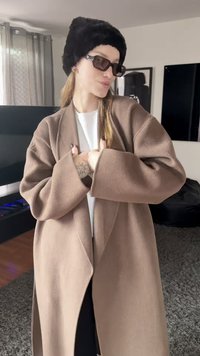 Manteau brun surdimensionné avec manches larges, tissu texturé, associé à un chapeau en fausse fourrure noir et des lunettes de soleil rectangulaires foncées.