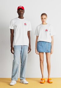 Hvite t-skjorter med kirsebærgrafikk, lys blå jeans, et denimskaft, og oransje sandaler. En bemerkelsesverdig rød caps på en modell. Enkel bakgrunn.