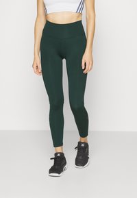 Gröna högmidjade leggings gjorda av elastiskt material med en mjuk textur, prydda med logotyputskrifter längs sidan, ihop med svarta sneakers.