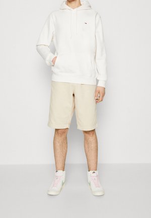 Bílá bavlněná mikina s kapucí a přední kapsou, béžové kraťasy a bílé vysoké tenisky s pastelovými akcenty, vystavené na neutrálním pozadí.