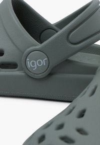Graue Kunststoffsandalen mit strukturierten Oberflächen, verstellbarem Riemen und kreisförmigem Logo. Auffällige Aussparungen für Belüftung und Flexibilität.