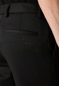 Black trousers with a smooth texture, featuring a back pocket, embossed "Porsche" and "BOSS" logos, and a sleek design.