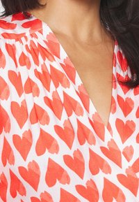 Blouse à col en V en tissu blanc avec un motif de cœurs rouges, texture douce et détail froncé à l'épaule.