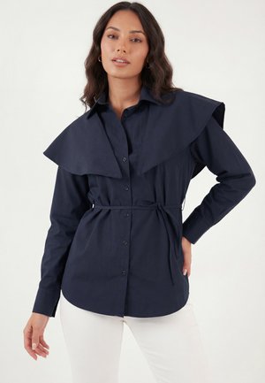 Nő sötétkék gombos blúzban, nagy gallérfülekkel és derékövtűzve, fehér nadrággal párosítva, egy kézzel a csípőn pózol.