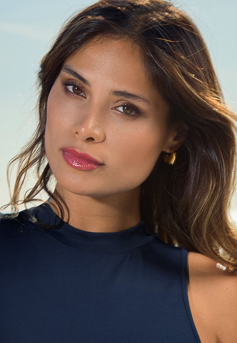 Gros plan sur une femme aux cheveux bruns ondulés, portant un haut marin col montant, un rouge à lèvres rose brillant et des boucles d'oreilles en or contre un fond lumineux.