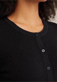 Svart texturerad kavaj med rund halsringning och fem svarta knappar. Tyget har ett upphöjt mönster som skapar visuell intress. 