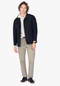 Chaqueta navy con cuatro bolsillos frontales, cuello abotonado, llevada sobre un polo gris claro. Pantalones cargo color beige claro y zapatillas negras con suelas marrones.