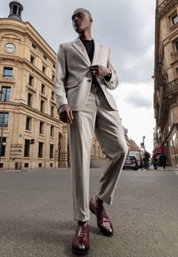 Completo grigio chiaro con vestibilità slim, giacca con due bottoni, abbinata a scarpe in pelle marrone scuro. Ambientazione urbana.