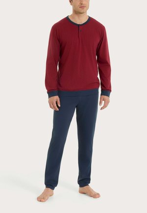 Uomo in piedi a piedi nudi che indossa una maglia rossa a maniche lunghe con polsini e colletto blu navy, abbinata a pantaloni blu navy, su uno sfondo chiaro uniforme.