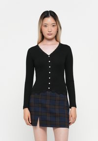 Cardigan noir à côtes avec des manches longues, col en V, bordure en dentelle festonnée, fermeture à boutons, associé à une mini-jupe à carreaux dans des tons de bleu foncé et de marron.